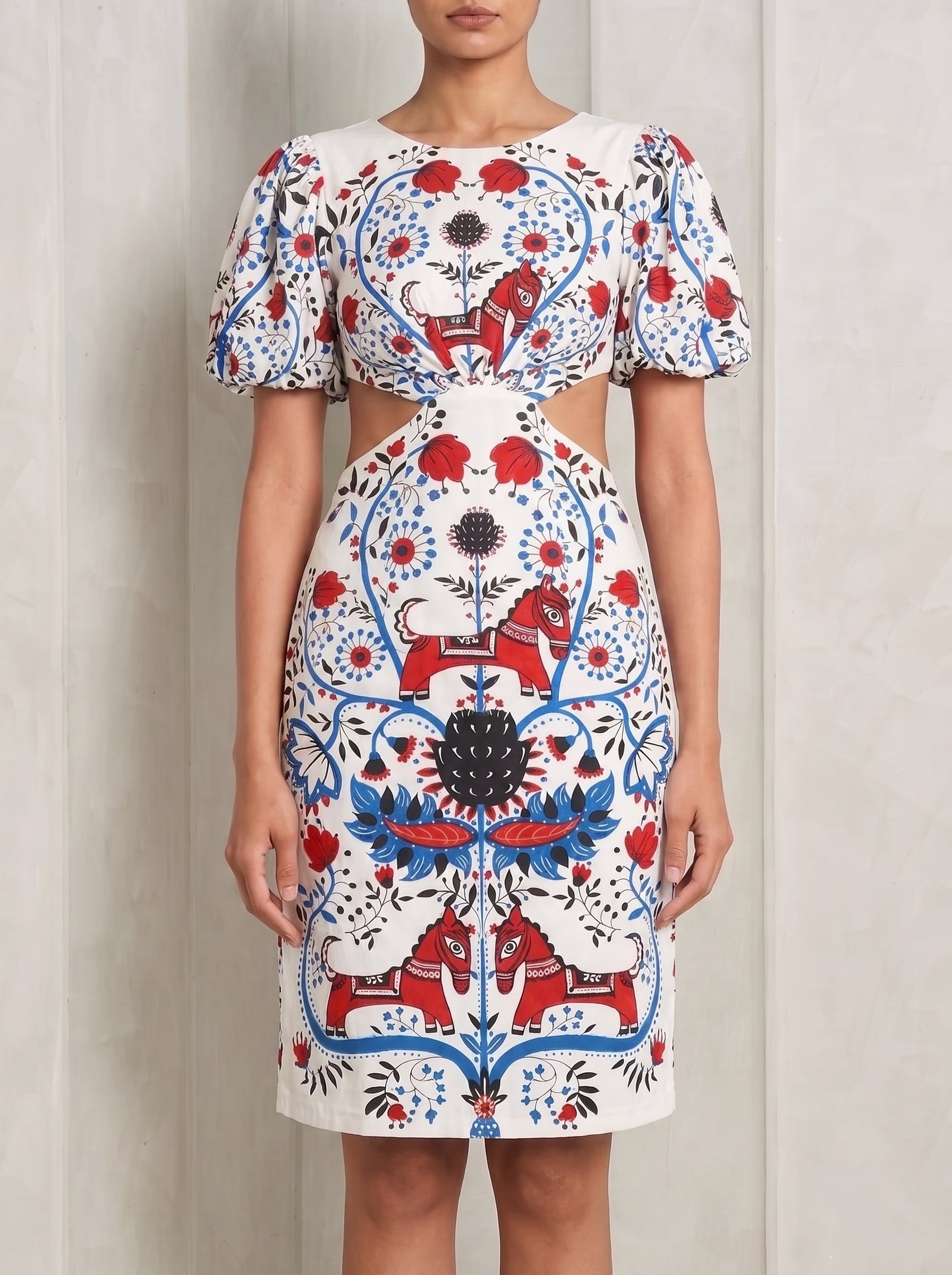 A woman modeling a fitted white dress with puff sleeves, a round neckline, and side cutouts at the waist. The dress features a bold folk-art style print in red, blue, and black with floral and horse motifs, set against a light neutral background.