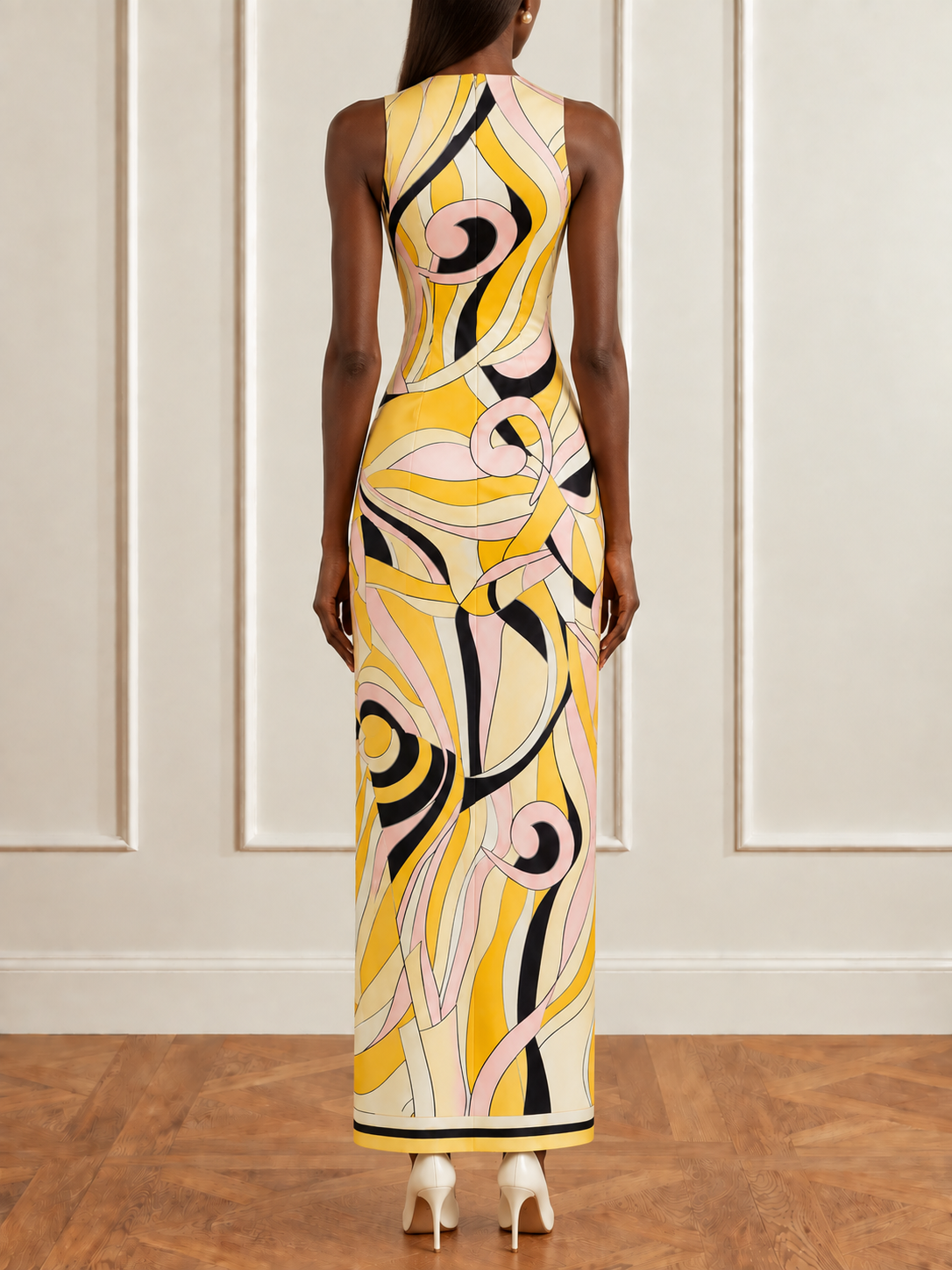 A woman viewed from the back wearing a sleeveless, floor-length dress with an abstract swirling pattern in yellow, cream, black, and light pink, standing on a wood floor against a paneled cream wall.